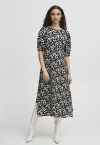 Robe noire à fleurs avec des manches bouffantes, présentant des motifs floraux légers, un col rond et une coupe fluide. Portée avec des bottes blanches jusqu'aux genoux.