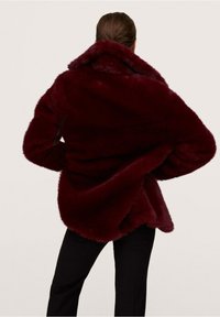 Mujer de pie con las manos en las caderas, vistiendo un abrigo de pelaje sintético burdeos oscuro y pantalones negros, contra un fondo claro y simple.