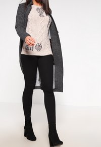 Cappotto a scacchi bianco e nero lungo su un maglione in maglia a treccia color crema, abbinato a leggings neri e stivali alla caviglia.