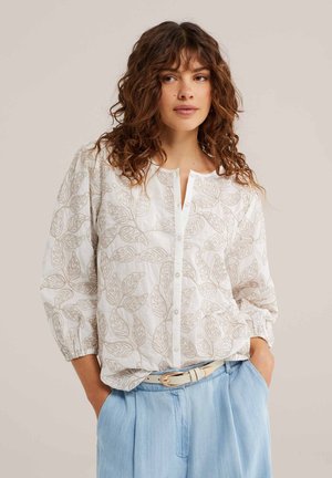 Femme aux cheveux bruns bouclés porte un chemisier blanc boutonné avec des motifs de feuilles beiges et un pantalon taille haute bleu clair avec une ceinture beige.