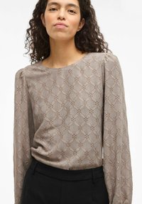 Ilgas smėlio spalvos viršus su tekstūruotu raštu su gėlių motyvais, apvalia iškirpte ir šiek tiek pufuotais pečiais. Juodos kelnės matomos juosmens srityje.