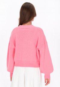 Ullig rosa tröja med en avslappnad passform, vida ärmar och ribbat fåll. Bakifrån syns en mjuk textur och en något kortad design.