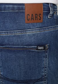 Jeans in denim con un'etichetta in pelle marrone con la scritta "CARS" impressa, una piccola etichetta nera sulla tasca, in tessuto blu, taglio dritto, con cuciture arancioni.