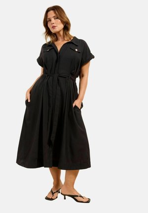 Naine, kes kannab musta midi pikkusega kleiti lühikeste rullitud varrukatega ja vööga taljes, seisab käed taskutes ja mustade kontsakingadega.