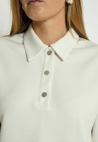 White polo shirt with a collar and five metallic buttons along the placket. Soft fabric texture with a clean, simple design.