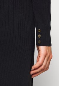 Czarna prążkowana dzianinowa sukienka z bocznym rozcięciem, ozdobiona trzema złotymi guzikami na mankiecie. Tkanina ma fakturowany wygląd.