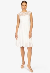 Robe blanche sans manches avec un corsage en dentelle et une jupe plissée fluide. Associée à des talons blancs à brides.