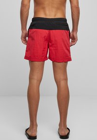 Røde badeshorts med svart elastisk midje, med en baklomme, glatt tekstur og midt på låret skjæring.