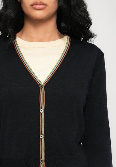 Cardigan blu navy con scollo a V, caratterizzato da una rifinitura a strisce multicolori e cinque bottoni grigi. Indossato sopra una maglia leggera color crema a coste.