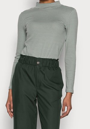 Zbliżenie osoby noszącej szarą i białą bluzkę w paski z długim rękawem oraz ciemnozielone, wysokie spodnie z zapięciem na guziki.