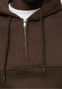 Felpa con zip marrone realizzata in tessuto morbido, dotata di tasca a marsupio, cerniera argento e cappuccio con cordoncino e estremità metalliche.