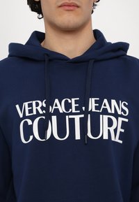 Marineblå hettegenser laget av bomull med snor; stor hvit tekst "VERSACE JEANS COUTURE" foran. Glatt tekstur.