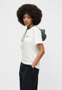 Γυναίκα με αφρο κόμμωση, που φορεί λευκό t-shirt Carhartt WIP και μαύρο παντελόνι, στέκεται με το ένα χέρι στην τσέπη, κοιτάζοντας πλάγια.