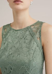 Grön spetsklänning med intrikata blommönster, ärmlös design, texturerad yta och vågig halsringning.