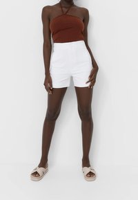 Vrouw in een bruine haltertop, witte high-waisted short en beige slipper sandaaltjes, staand tegen een witte achtergrond.