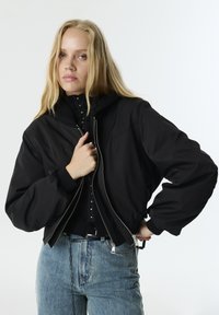 Zwarte bomberjack met een ritssluiting aan de voorkant, ribgebreide manchetten en hoge kraag. Naadloos ontwerp met een glad, waterafstotend materiaal. Casual cropped stijl.