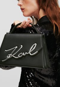Borsa a mano in pelle nera con una forma strutturata, caratterizzata da un piccolo logo "Karl" in argento ben visibile. Dotata di un manico corto per il trasporto a mano.