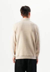 Beige fleece pullover med høj krave, der viser en tekstureret overflade, ribbede manchetter og en afslappet pasform. Bagfra set med mørke bukser.