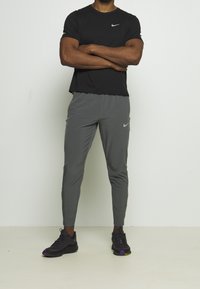 Man standing with arms crossed wearing black Nike t-shirt, gray Nike track pants, and black sneakers with purple and yellow accents.