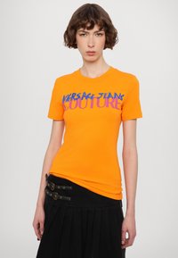 Oranžové bavlněné tričko s textem "VERSACE JEANS COUTURE" v modré a růžové barvě. Doplněno černou sukní s sklady a detailně s přezkou.