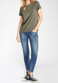 Camiseta de algodón verde oliva con diseño estampado, combinada con jeans ajustados de mezclilla azul y zapatillas negras de caña alta.