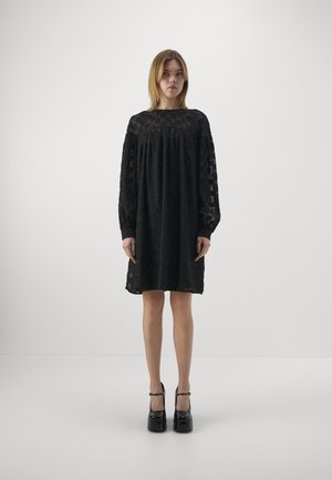 Vestido negro texturizado con mangas largas, escote redondo y cintura fruncida. Presenta un patrón sutil y cae por encima de las rodillas. Combinado con tacones de plataforma.