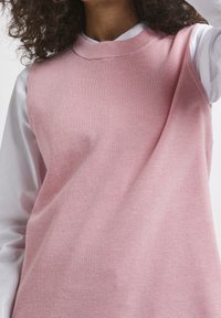 Top senza maniche lavorato a maglia rosa con una texture a costine e un collo rotondo, abbinato a una camicia bianca con colletto a maniche lunghe sotto.