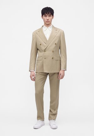 Uomo che indossa un abito beige doppiopetto con camicia bianca e sneakers bianche, in piedi davanti a uno sfondo bianco uniforme.
