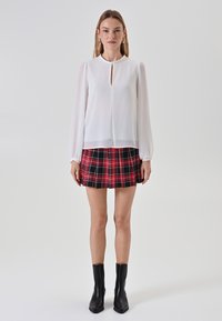 Weiße Bluse mit langen, gepufften Ärmeln und einem Schlüssel-Ausschnitt, kombiniert mit einem schwarz-roten karierten, gefältelten Minirock. Schwarze Knöchelstiefel.
