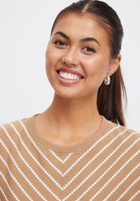 Pull marron avec des rayures en chevron blanches, col rond et tricot texturé. Modèle portant des boucles d'oreilles en argent, souriant, en plan rapproché.