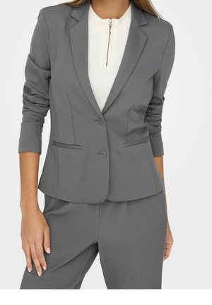 Femme portant un blazer gris à deux boutons sur un haut blanc à fermeture éclair et un pantalon gris assorti, debout avec une main près de sa cuisse.