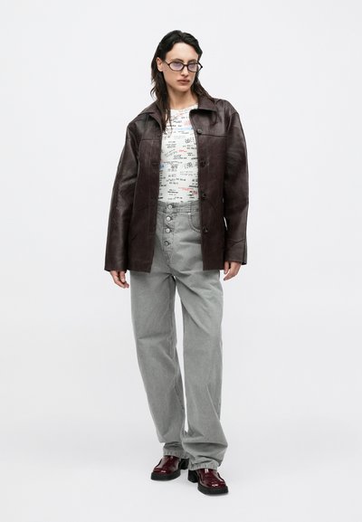 Bruine leren jas met een gestructureerd ontwerp, wit T-shirt met afbeelding, grijze broek met wijde pijpen en glanzende bordeauxrode schoenen.