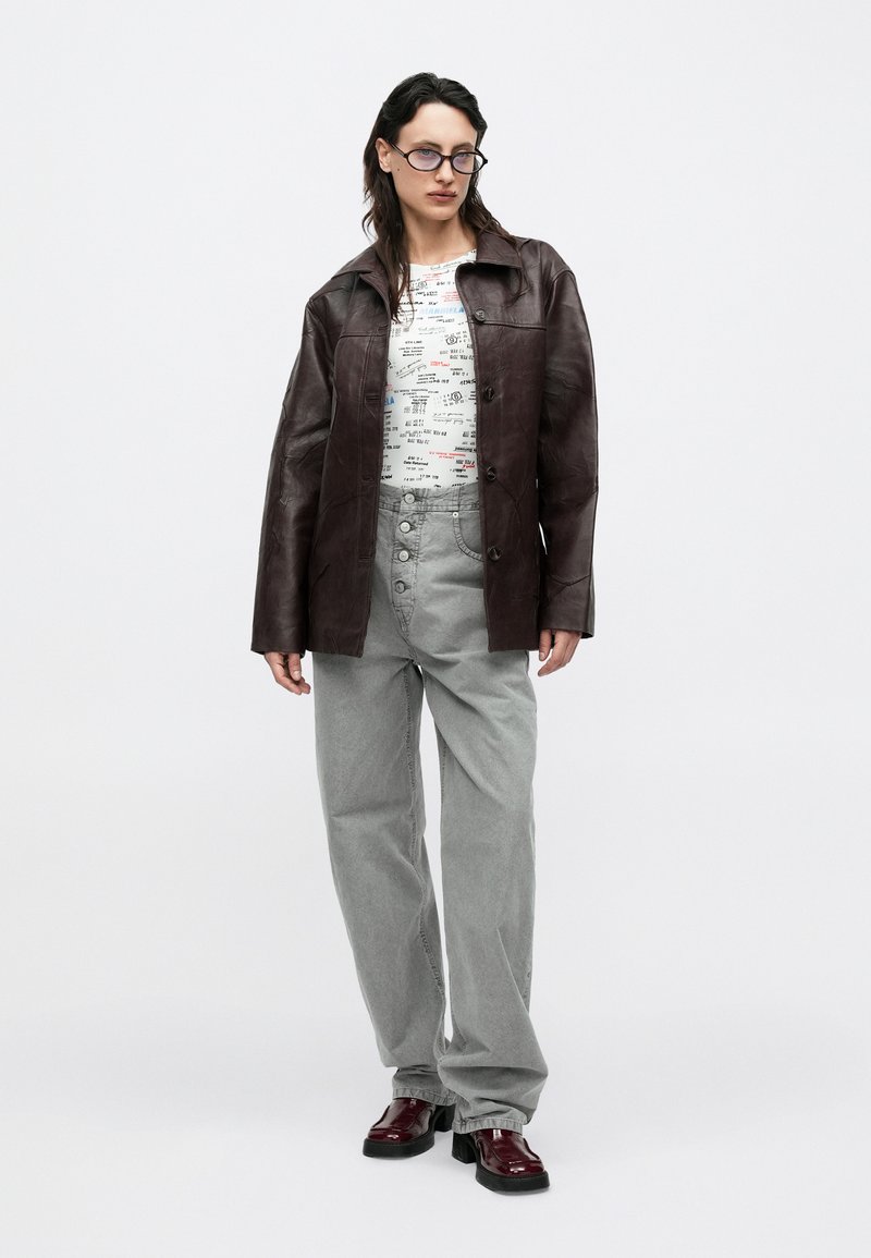 Veste en cuir marron au design structuré, t-shirt blanc à motif graphique, pantalon gris à jambes larges, et chaussures bordeaux brillantes.