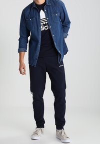 Jeansová košile na knoflíky přes černé tričko Adidas, doplněná námořnickými tepláky s bílými pruhy. Celkový vzhled doplňují neutrální tenisky.