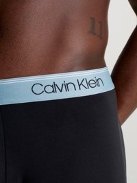 Schwarze Boxer mit einem hellblauen elastischen Bund, der das Logo "Calvin Klein" in dunkler Schrift zeigt. Glatte Stoffoberfläche, körpernahe Passform.