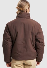 Brun puffer-jakke med høy krage, med en matt tekstur og synlige sømmer. Løs design med ribbede mansjetter på ermene.