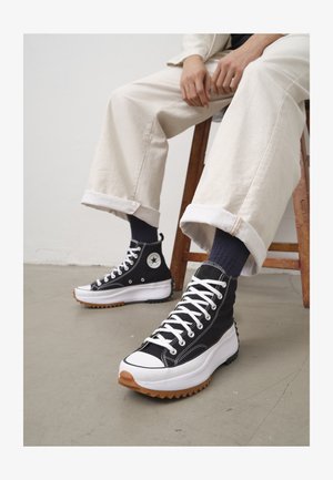 High-top black sneakers with white laces and rubber toe cap. Thick white sole with a textured brown outsole, worn with navy striped socks.