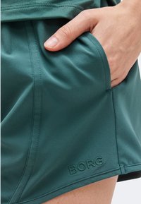 Χέρι που ακουμπά στην τσέπη των τιρκουάζ αθλητικών σορτς με ορατές γραμμές ραφής και το λογότυπο «BORG» κοντά στο τελείωμα.