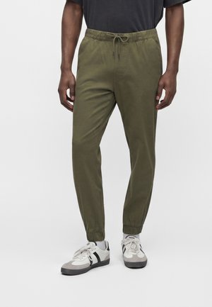 Homme portant un pantalon de jogging vert olive avec des ourlets élastiqués et des baskets blanches avec des accents noirs, debout sur un fond uni.