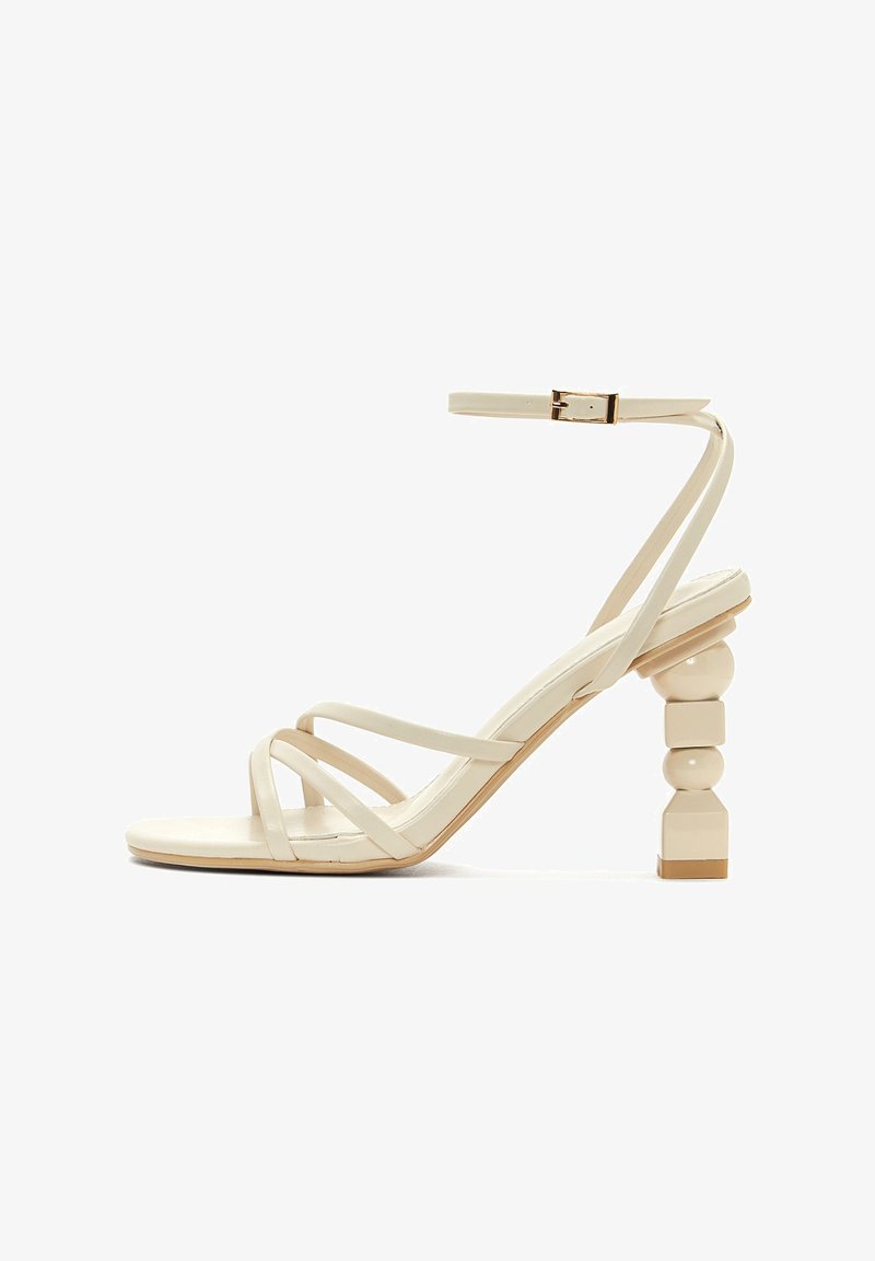 Sandale beige à brides avec un talon carré, ornée de formes rondes et carrées, en matériau verni et dotée d'une sangle pour la cheville avec une boucle.