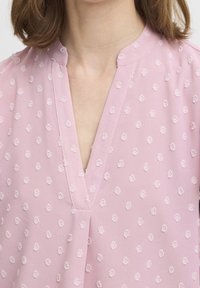 Jasnoróżowa bluzka z dekoltem w kształcie litery V, ozdobiona teksturowanymi kwiatowymi wzorami na całej powierzchni. Gładka tkanina z delikatnym połyskiem i subtelnymi zdobieniami.