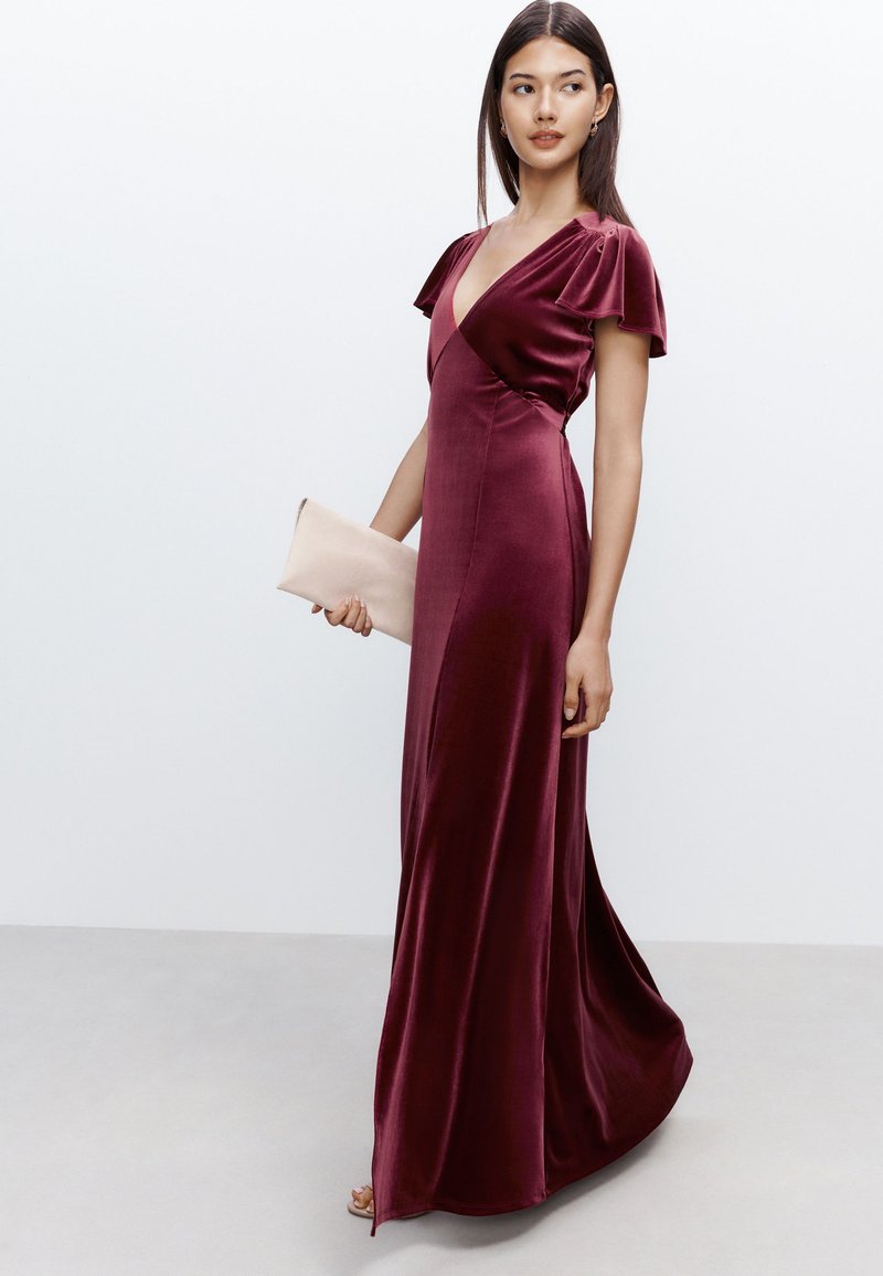 Robe longue en velours bordeaux avec un col en V profond, des manches volantées et une silhouette fluide. Le mannequin tient une pochette rose clair.