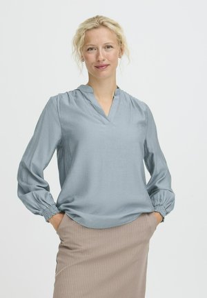 Himmelbluse mit V-Ausschnitt, langen Ärmeln, die an den Bündchen gerafft sind, aus glattem Stoff. Kombiniert mit einem beigen Nadelstreifenrock.