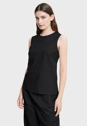 Młoda kobieta z prostymi brązowymi włosami, ubrana w czarny top bez rękawów i czarne spodnie na jednolitym tle.