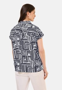 Blusa a maniche corte di colore bianco con un motivo geometrico nero. Il materiale sembra leggero con una vestibilità comoda e un dettaglio di piega sul retro.