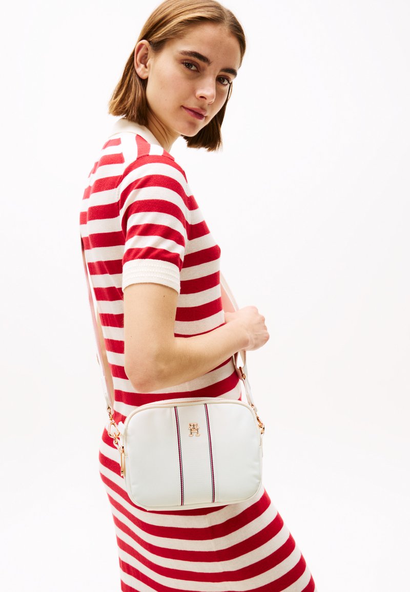 Borsa a tracolla bianca con strisce viola e bianche, accessori dorati e texture morbida, indossata da un modello in un abito a strisce rosse e bianche.