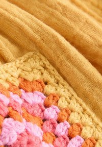 Un tissu en lin jaune drape un carré en crochet avec un motif texturé dans des tons de rose, d'orange et de jaune.