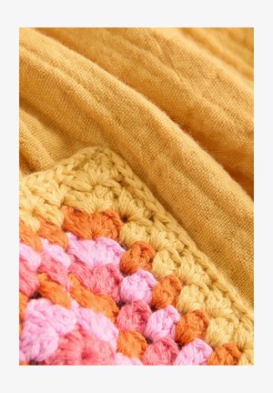 Žlutá lněná látka visí přes háčkovaný čtverec s texturovaným vzorem v odstínech růžové, oranžové a žluté příze.