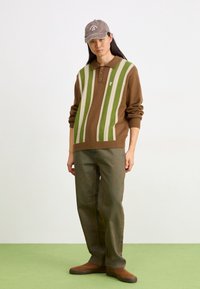 Jeune personne portant une casquette marron, un pull marron avec des rayures verticales vertes et crème, un pantalon marron ample et des chaussures marron, debout sur un sol vert.