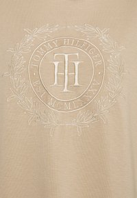 T-shirt beige con un logo circolare ricamato che presenta il testo "Tommy Hilfiger" e "EST. MCMLXXXV", circondato da un design di alloro.
